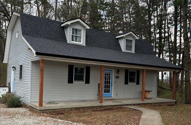 Small white house with a black roof and front porch in a wooded setting