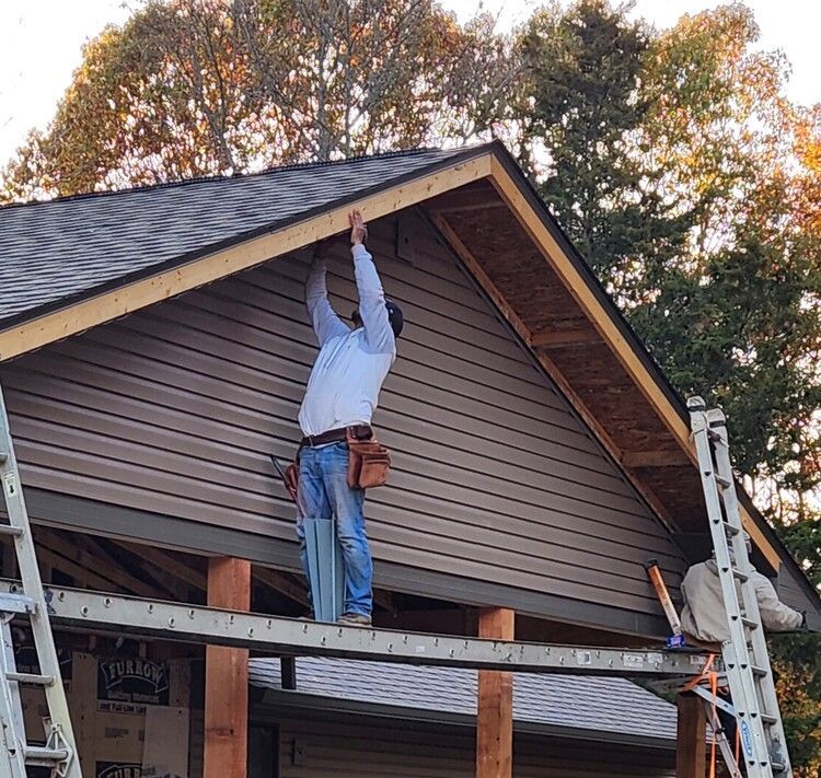 Worker standing on scaffolding reaching up to install siding under a house roof eave