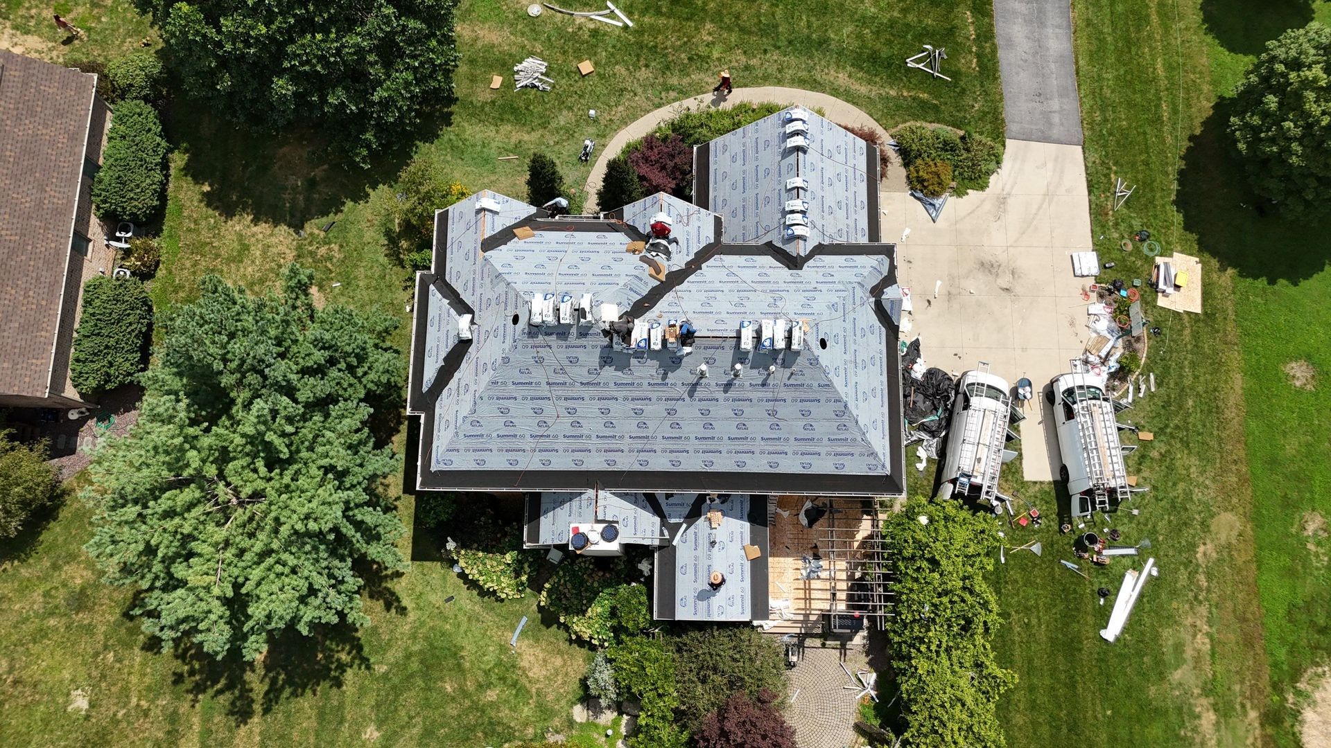 Aerial view of a house with a gray roof, surrounded by green trees and lawn, with debris and a vehicle at the driveway.