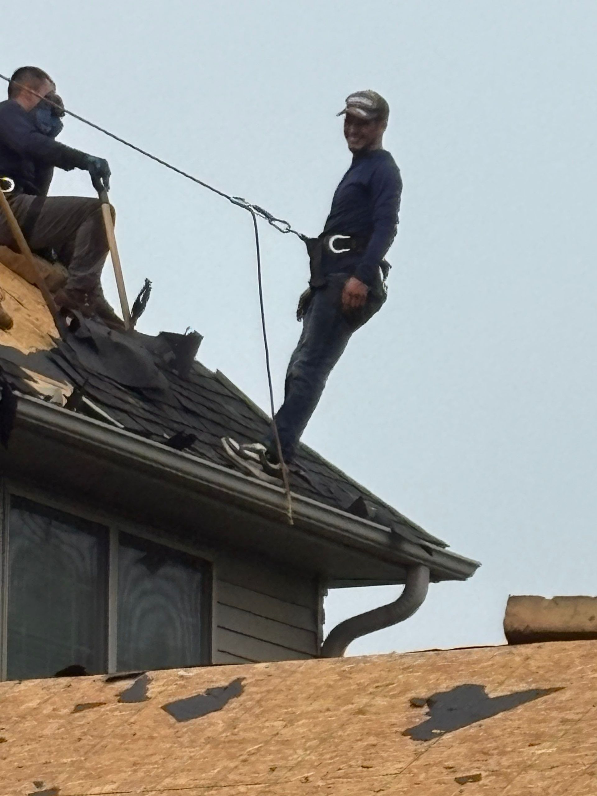 Two workers standing on a damaged roof, one near a ladder, with dark shingles being repaired.