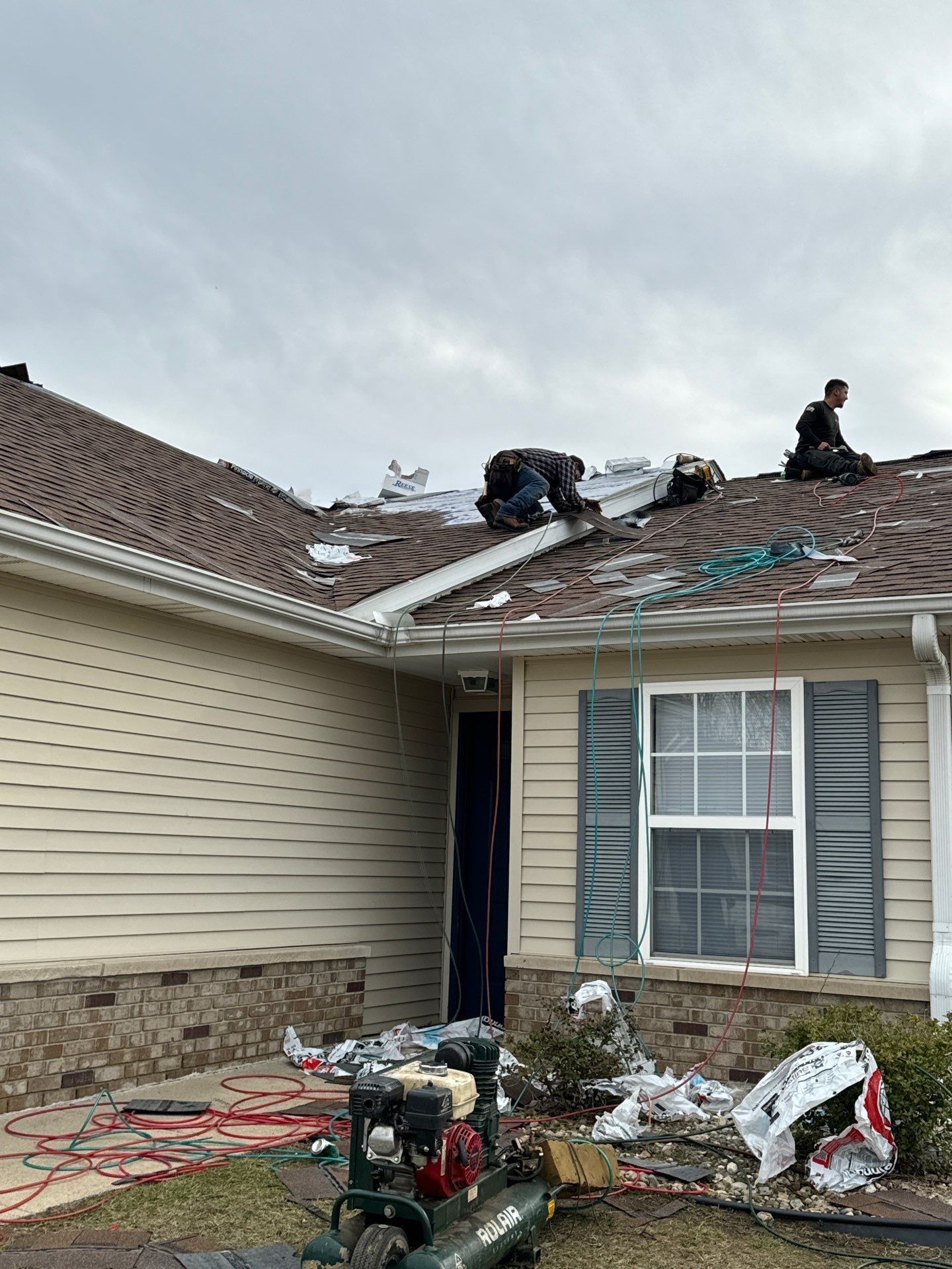 Workers repairing storm-damaged roof on a beige house, with debris and tools in the yard.