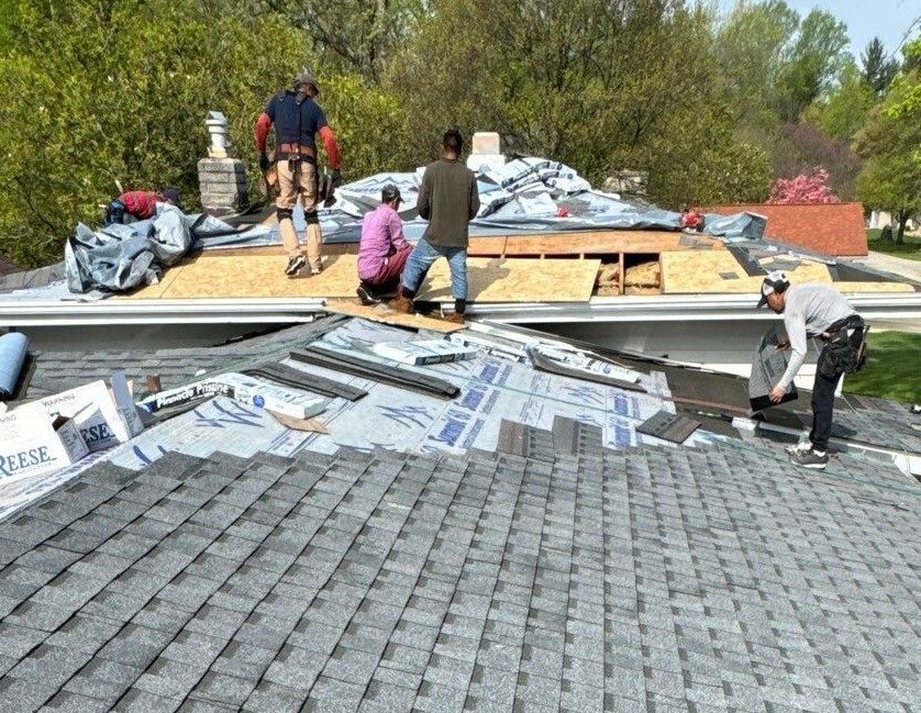 Workers installing roofing shingles on a house roof, with materials stacked nearby.
