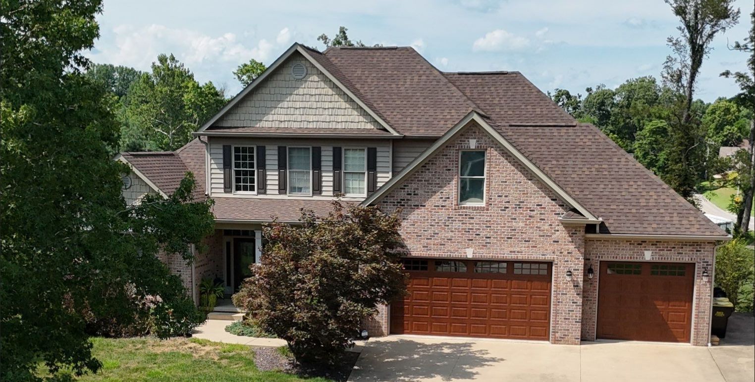 Two-story brick suburban house with a brown roof and three-car garage, surrounded by trees and a driveway