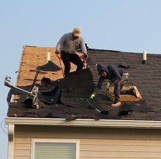 Two roofers removing damaged shingles from a house roof under a clear sky