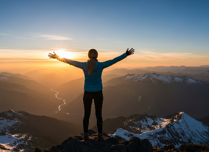 Person standing on a mountain peak at sunrise, arms outstretched over a valley and snowy ridges