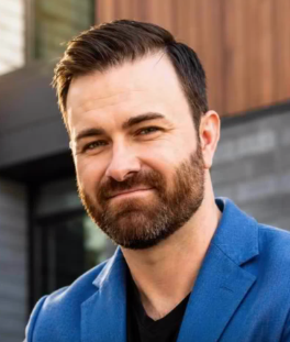 Man in a blue blazer and black shirt, smiling outdoors against a modern building.
