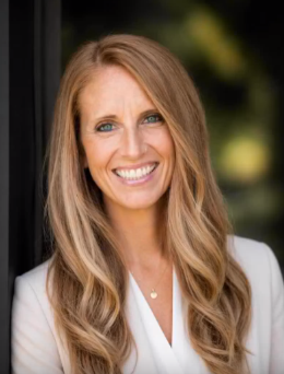 Smiling woman with long blonde hair in a white blazer against a blurred dark background