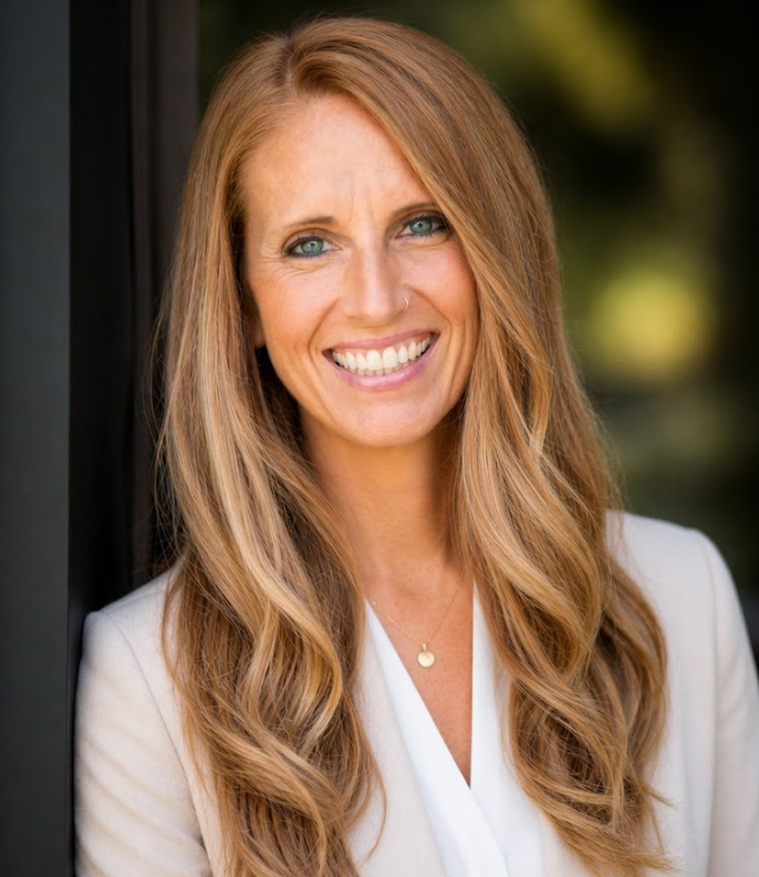 Smiling woman with long wavy blond hair in a white blazer against a blurred dark background