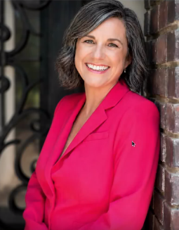 Smiling person in a bright pink blazer leaning against a brick wall