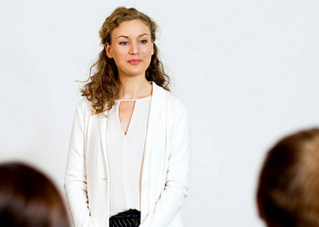 Woman in a white blazer standing at a podium against a plain white background