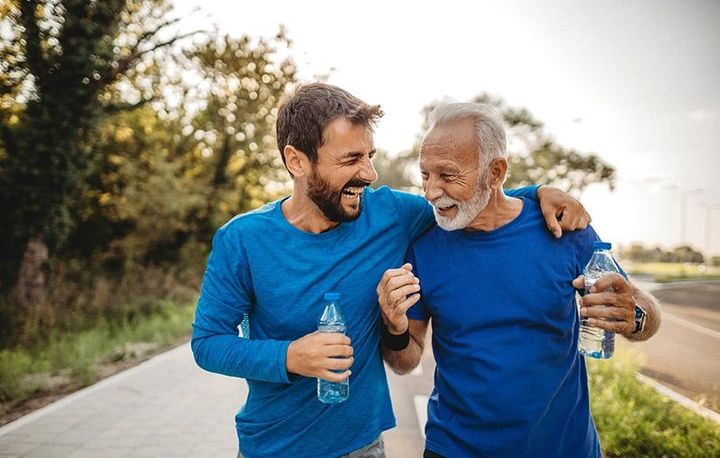 Two men in blue shirts smiling and walking outdoors with water bottles on a sunny path