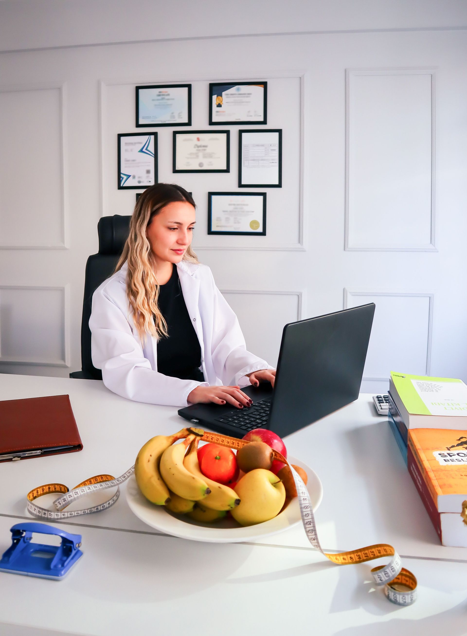 Woman typing on a laptop at a desk with fruit, notebooks, and framed documents on the wall.
