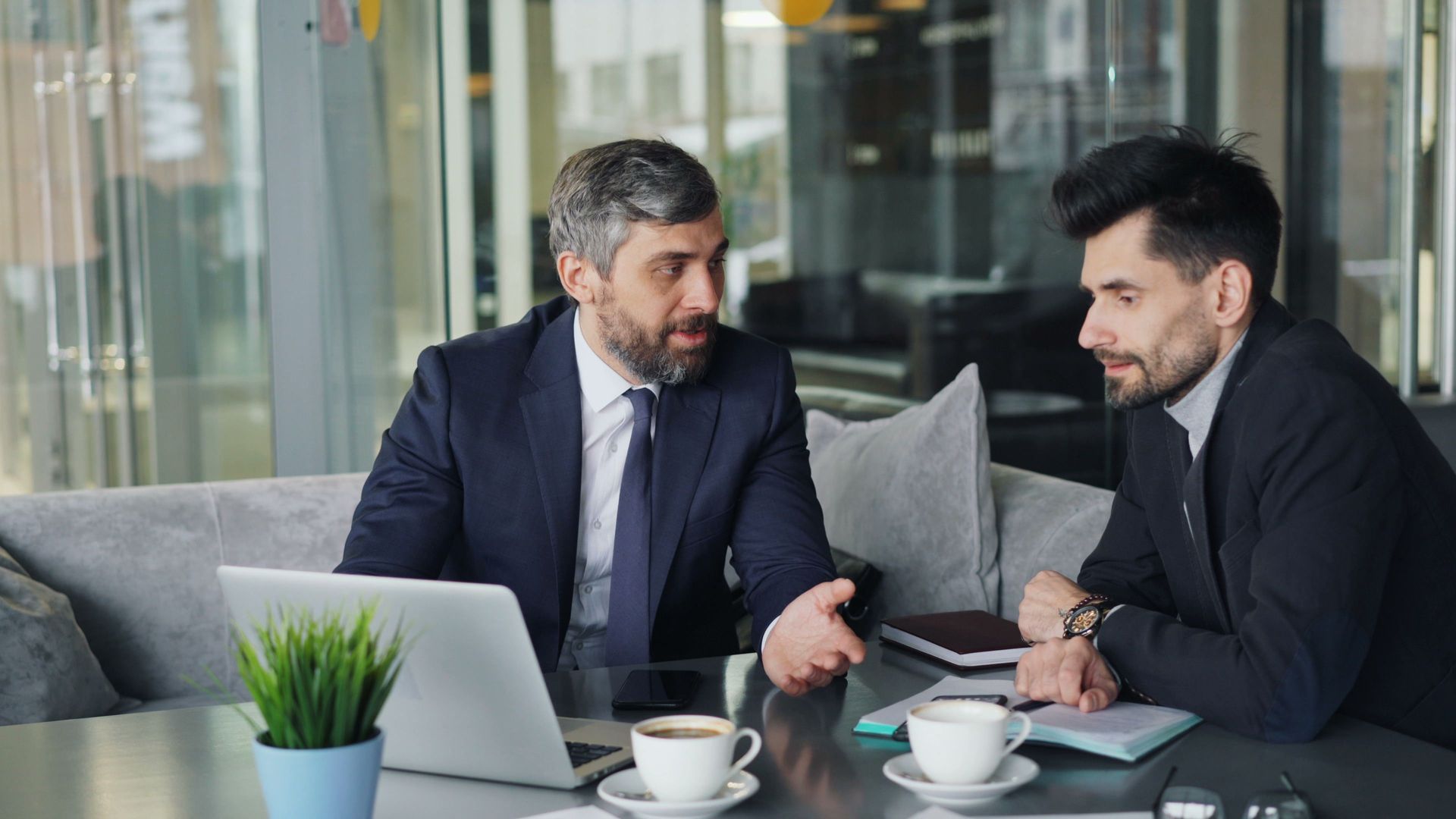 Two men in suits discuss business at a café table with a laptop and coffee cups.