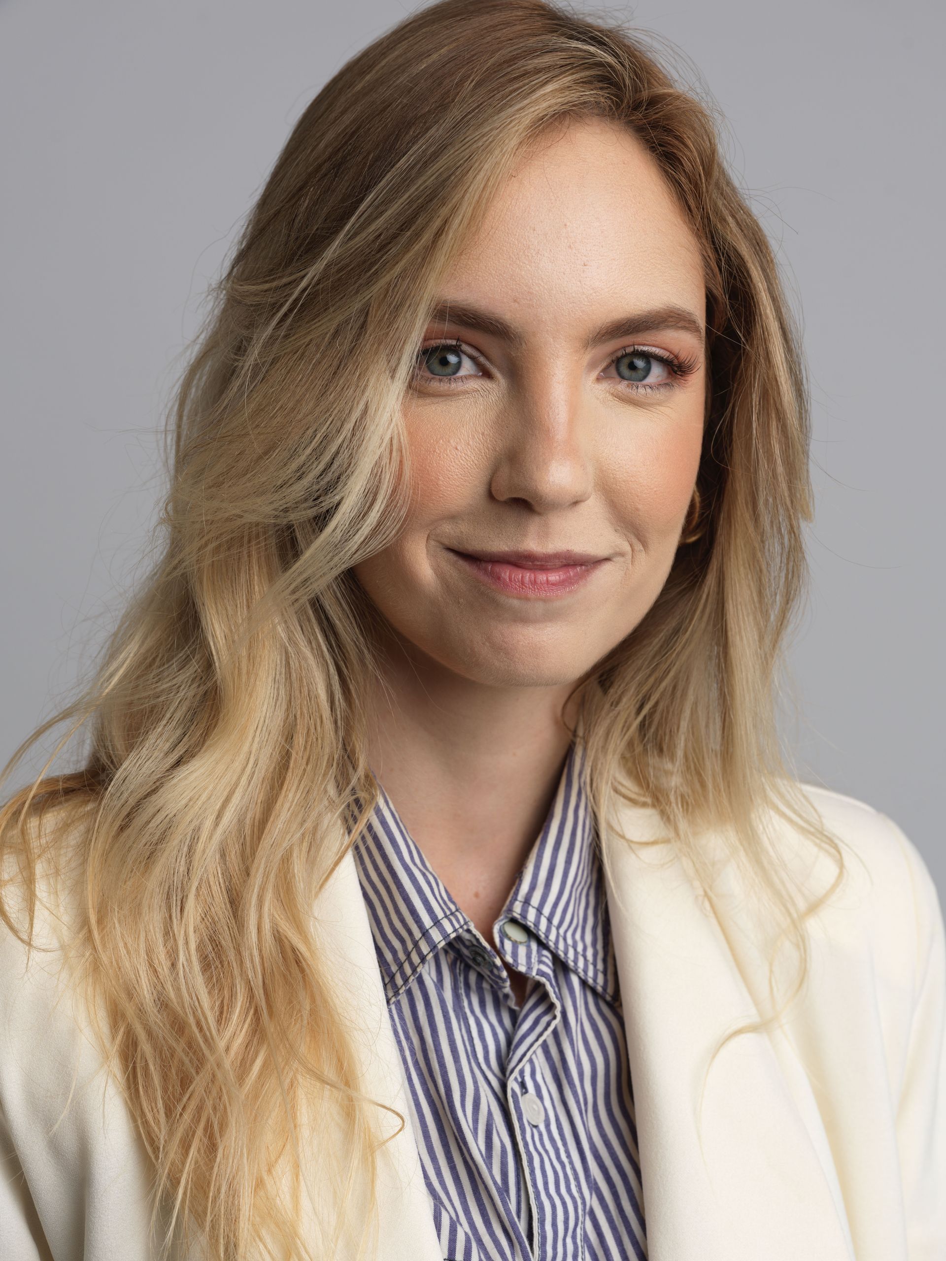Portrait of a smiling person in a white blazer and striped shirt against a gray background