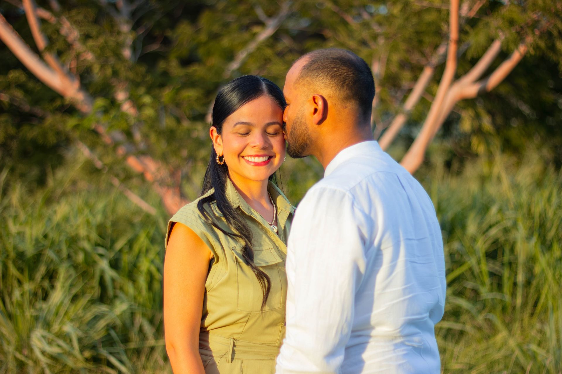 Couple standing close in a grassy outdoor setting, smiling in warm sunlight.