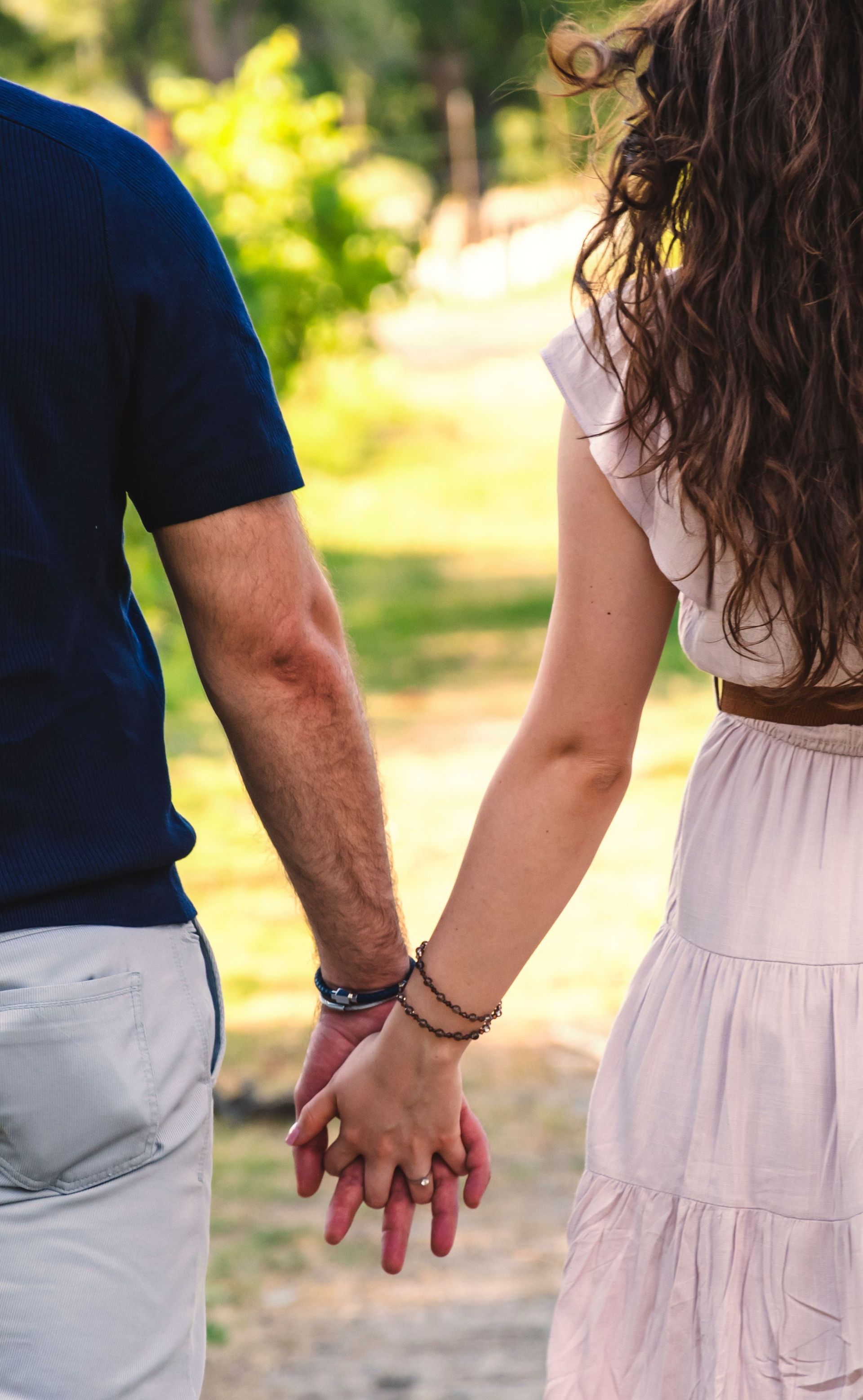 Two people holding hands while walking on a sunlit path, seen from behind