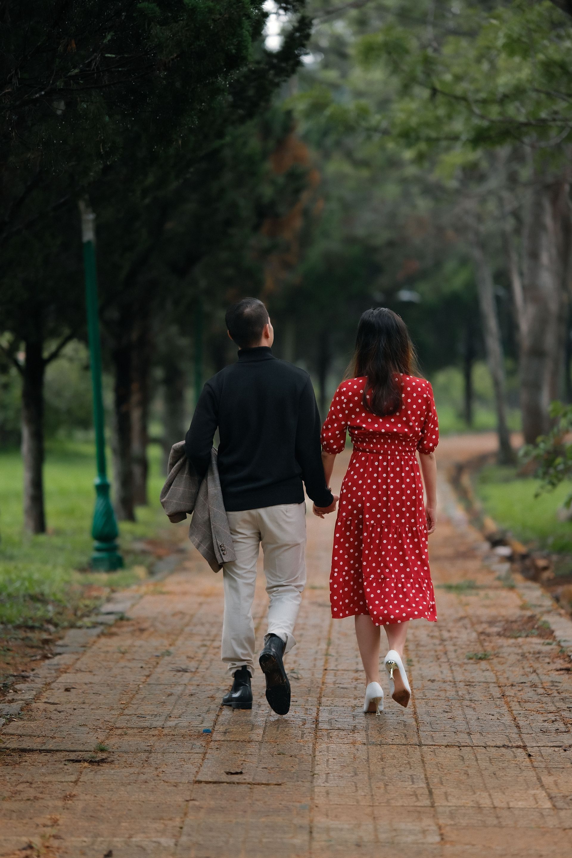 Two people walking away along a tree-lined path, one in black and beige, the other in a red polka-dot dress.
