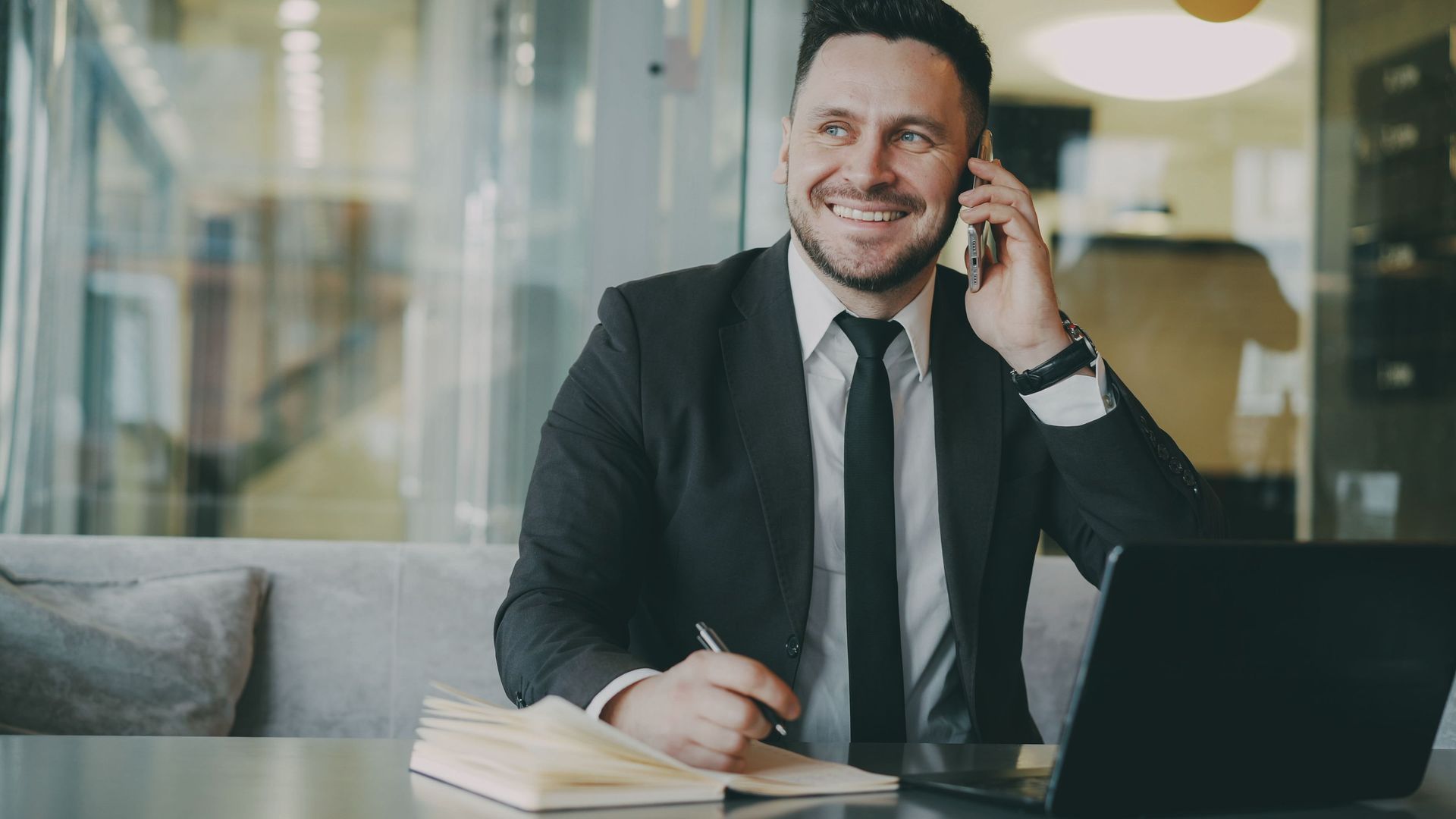 Man in a black suit smiling while on a phone call at a desk with a laptop in an office.
