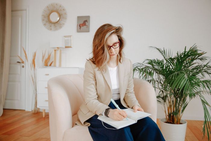 Woman writing in a notebook on a beige chair in a bright living room with plants and wall art