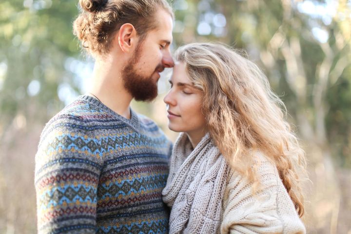Two people in sweaters leaning close outdoors, with soft trees in the background.