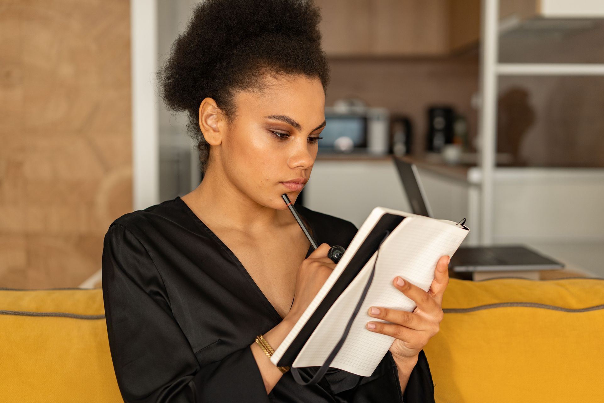 Person writing in a notebook on a yellow couch, with a laptop nearby.