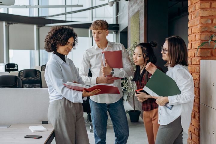 Four coworkers discuss documents in a modern office, standing by a brick wall and whiteboard.