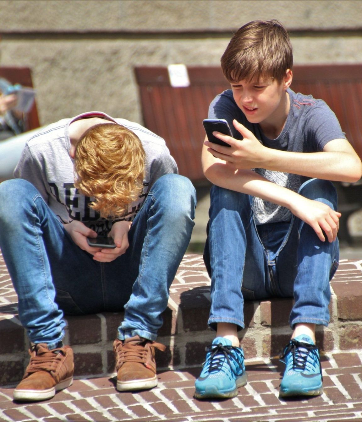Two boys are sitting on a brick sidewalk looking at their phones