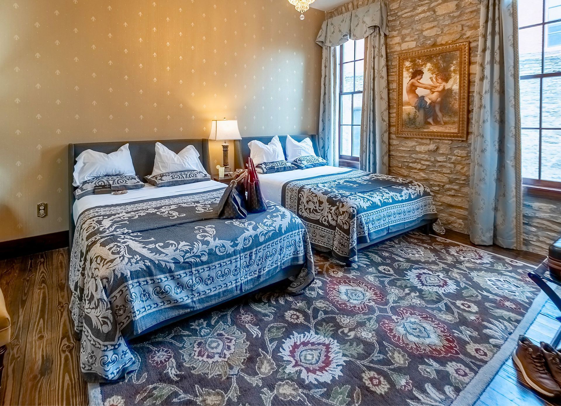 A bedroom with two beds, ornate blue bedding, a patterned rug, stone wall, and gold-framed art.