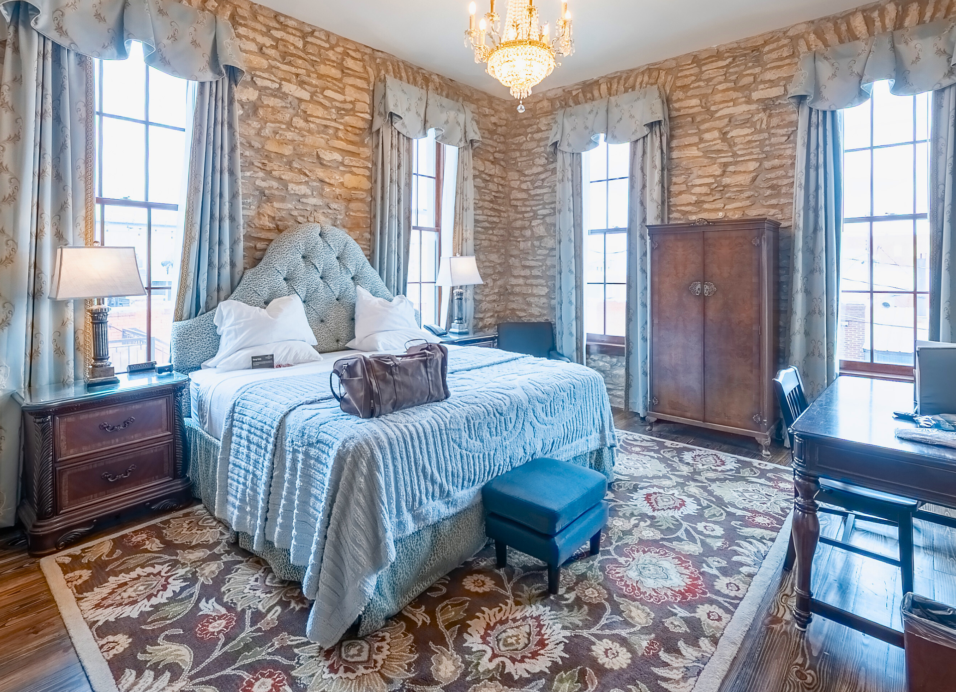 A luxury hotel bedroom with a large bed, patterned wallpaper, wooden furniture, and a crystal chandelier.