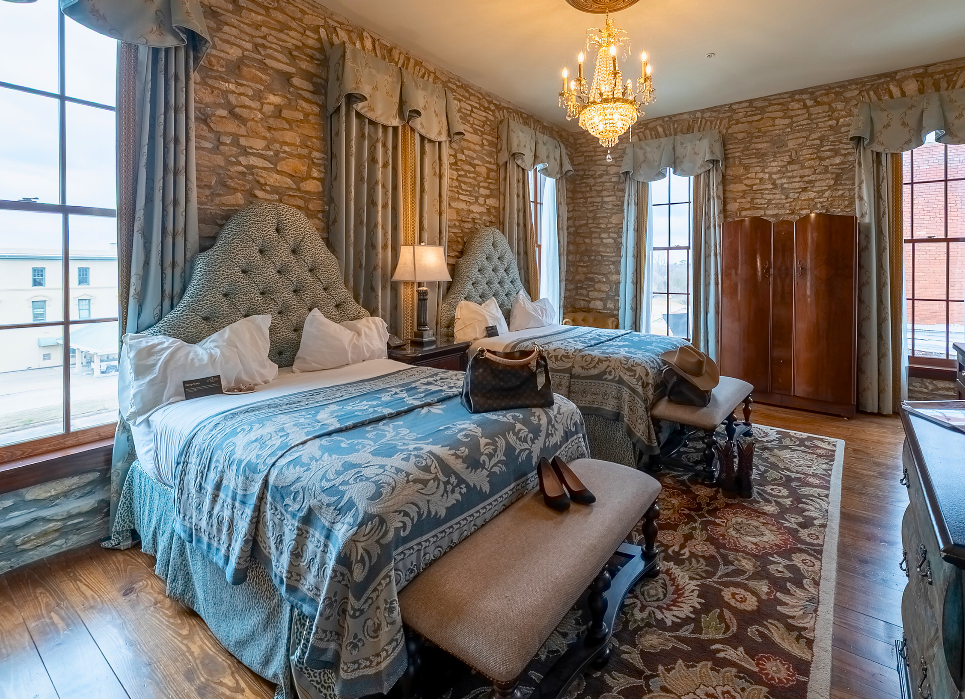 A bedroom featuring two beds with patterned blue bedding, textured wallpaper, heavy drapes, and a central chandelier.