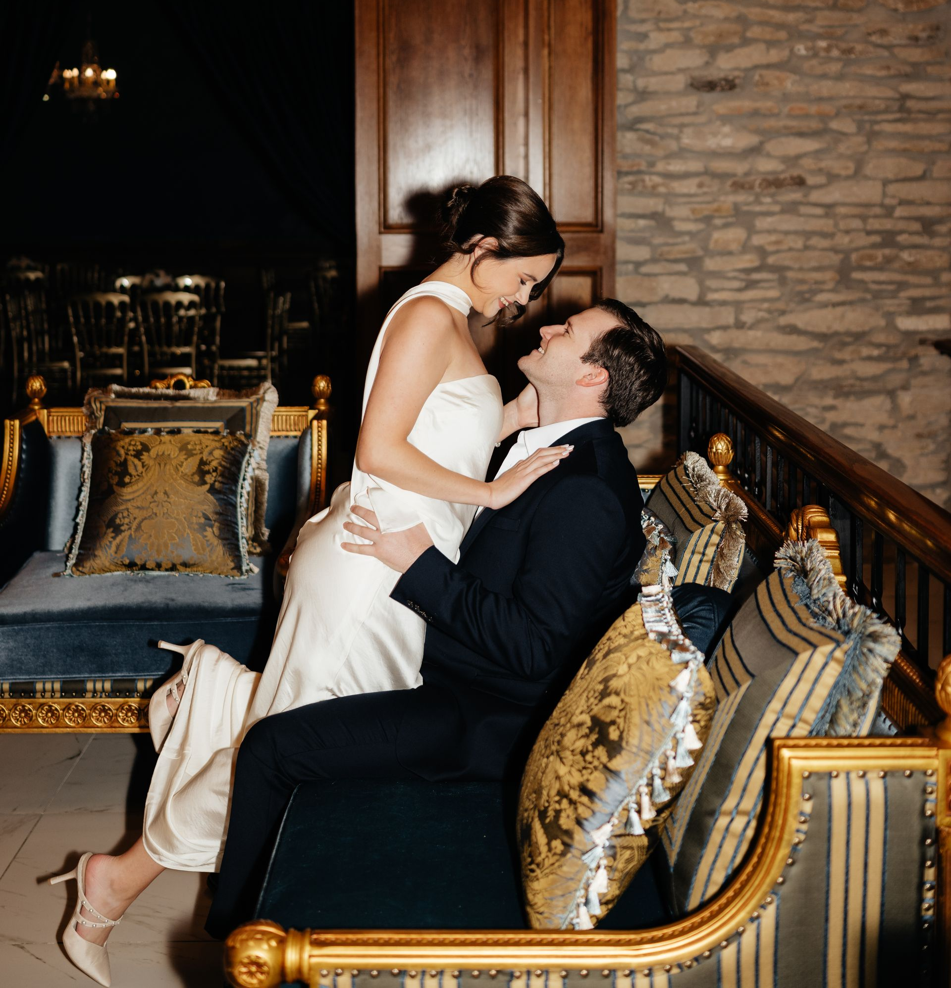 A person in a white gown sits on the lap of a person in a dark suit on a decorative sofa in a rustic, elegant room.
