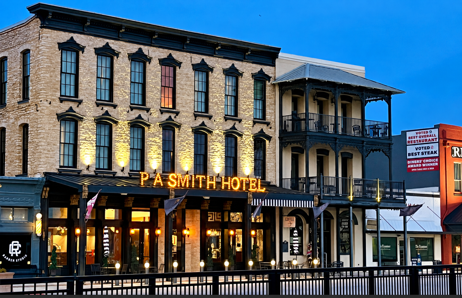 A three-story brick P.A. Smith Hotel building with a black balcony, lit against a deep blue dusk sky.