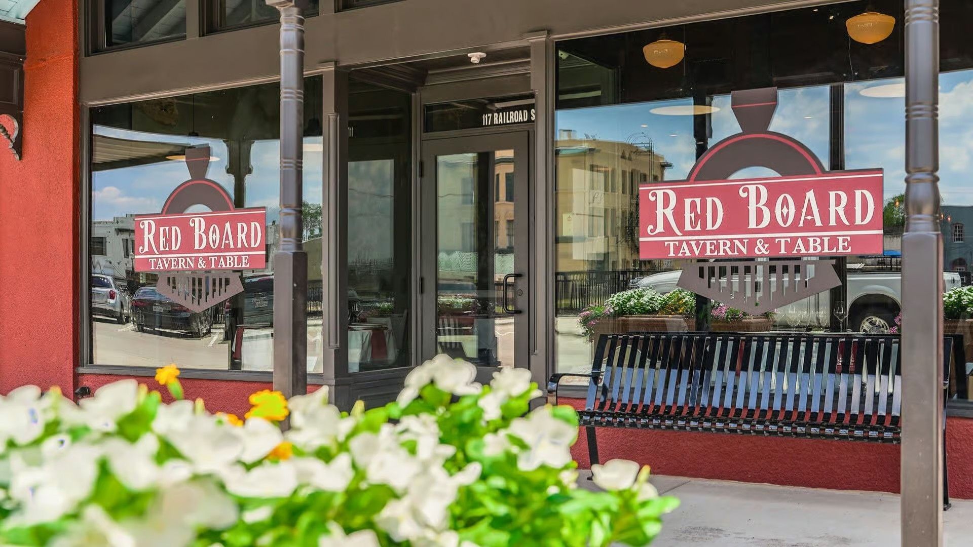 A storefront for Red Board Tavern & Table with red trim, glass windows featuring the logo, and flowers in the foreground.
