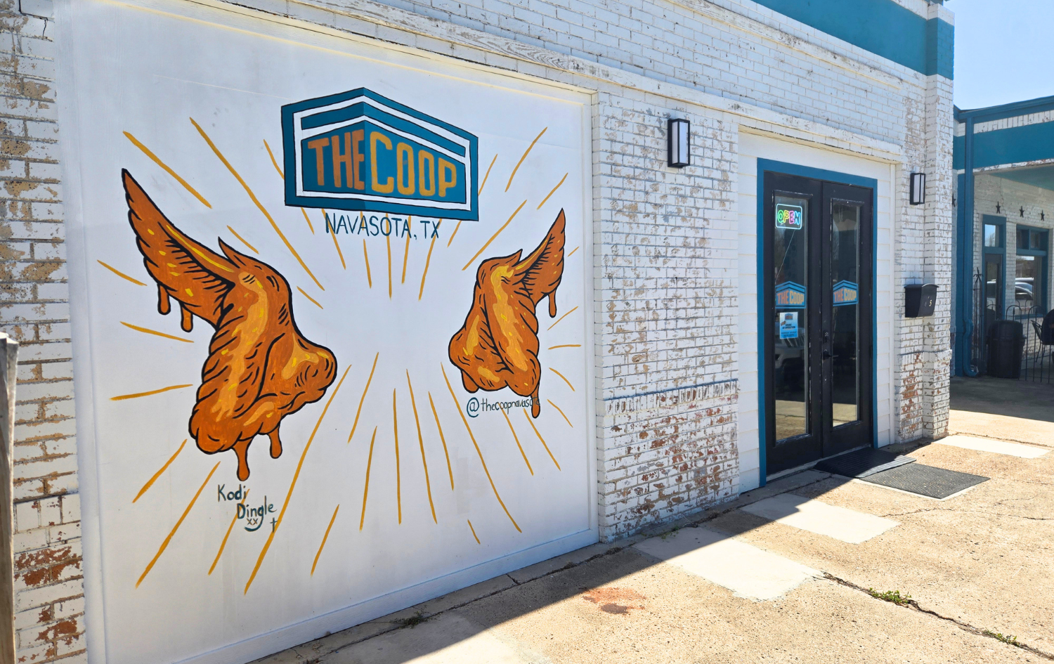 Mural for The Coop in Navasota, TX, featuring fried chicken wings as angel wings on a white brick restaurant exterior.