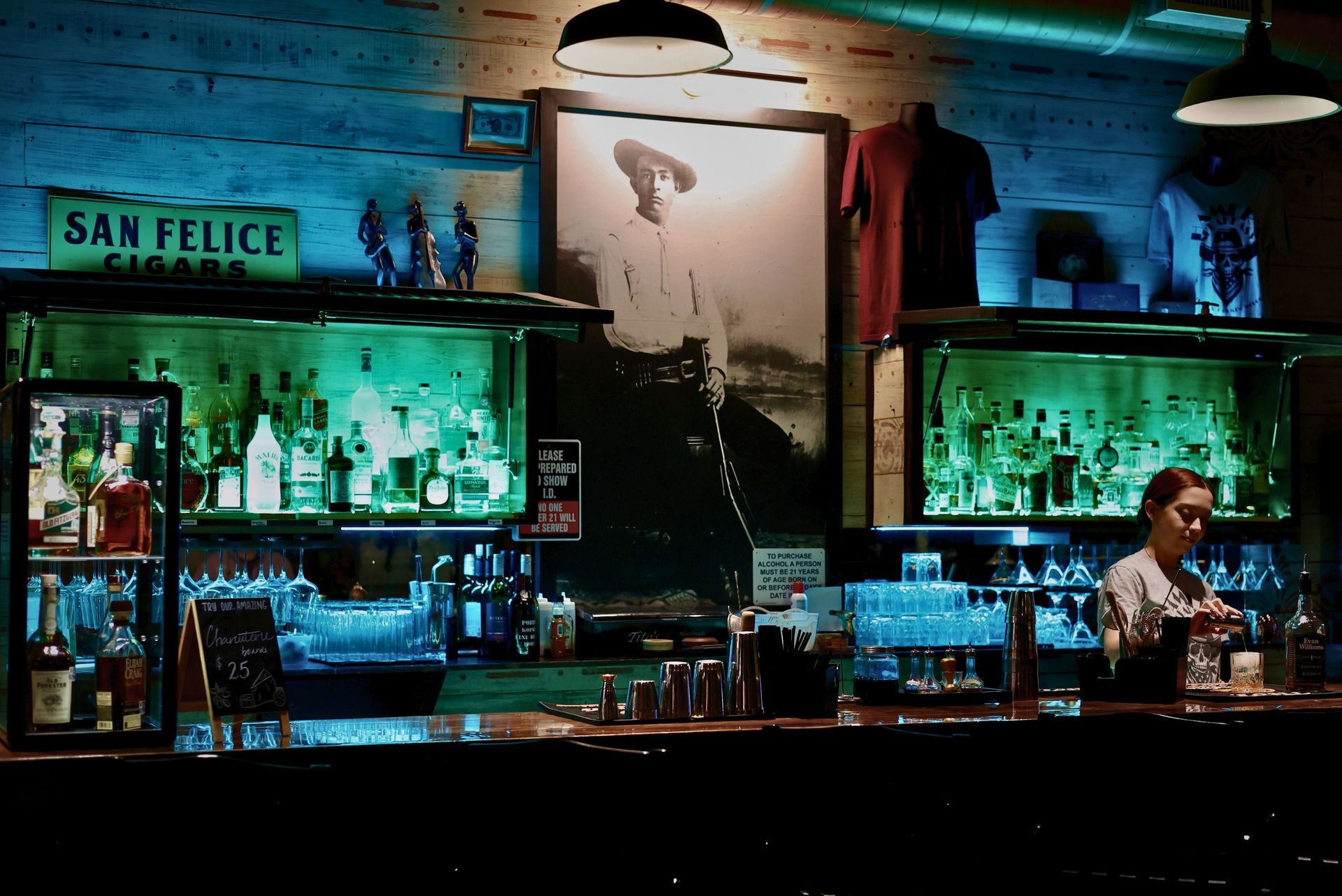 Bartender mixing drinks at a rustic bar with green-lit bottle shelves and a vintage cowboy portrait on a wood wall.