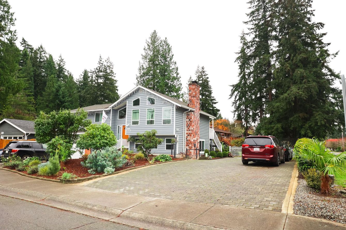 A Grace of Love Adult Family Home Gray house with brick chimney and a stone driveway, surrounded by greenery and trees.