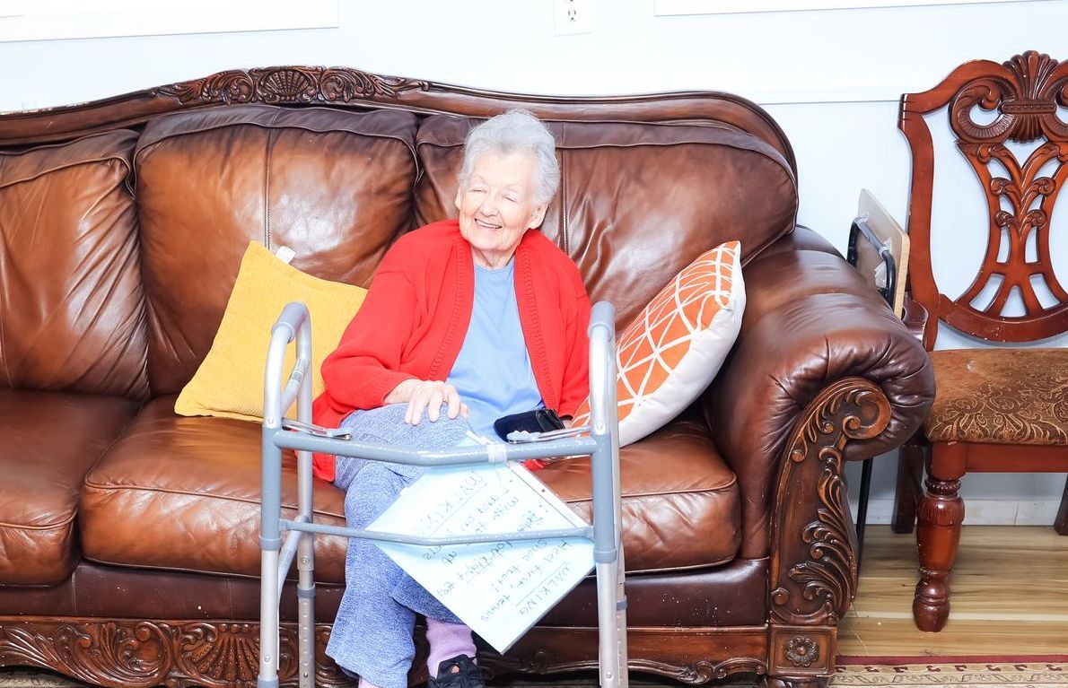 A Grace of Love Adult Family Home Elderly person smiles on a brown leather couch, holding a walker and papers in a home setting.