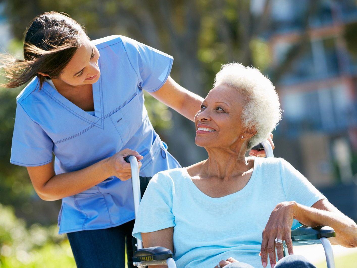Woman in wheelchair being pushed by caregiver outdoors.