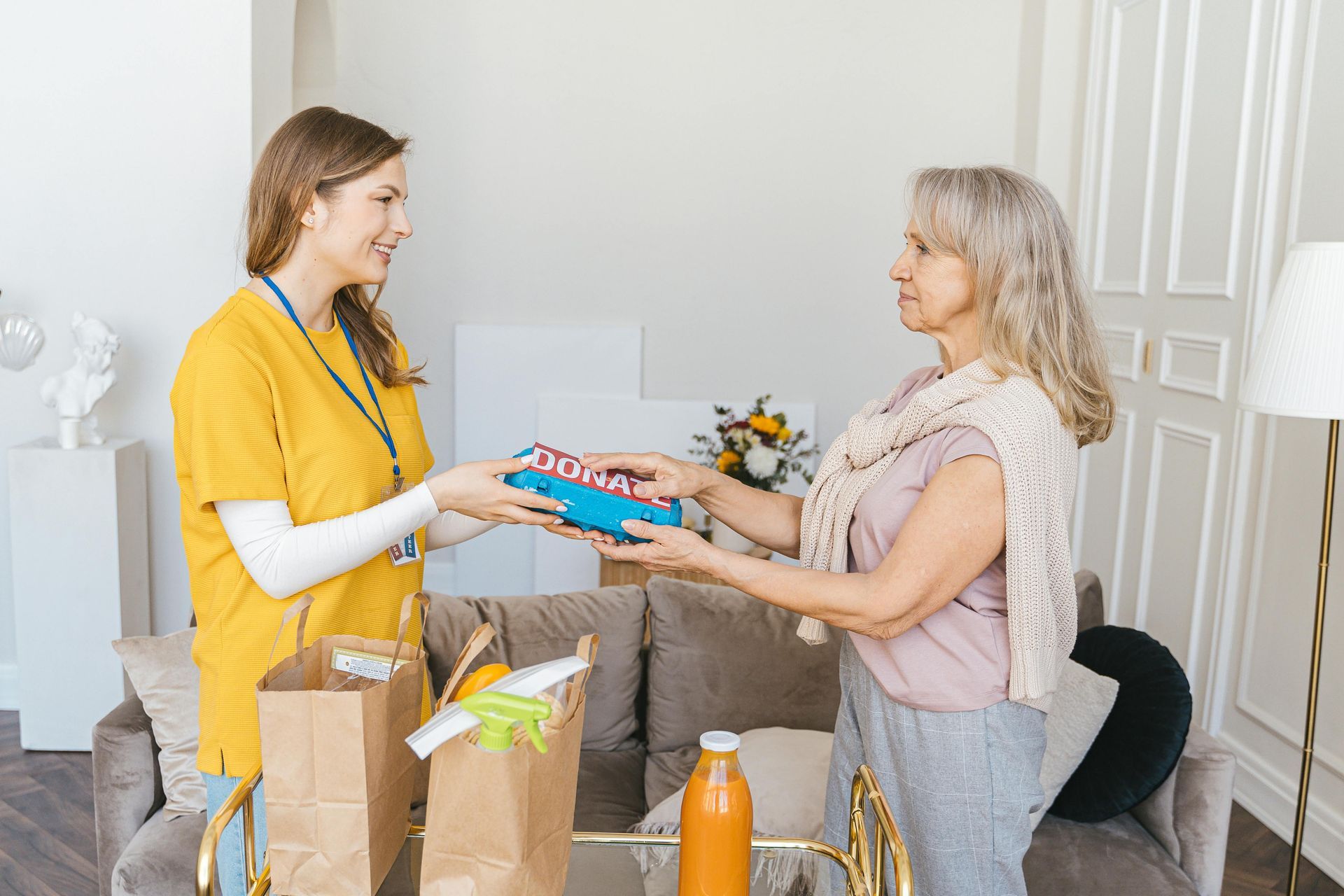 Woman in yellow shirt hands a blue box to another woman. Food bags on the sofa.