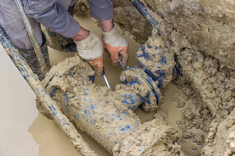 A Man is Working on a Muddy Pipe in the Dirt — De Rose Plumbing In Smithfield, QLD