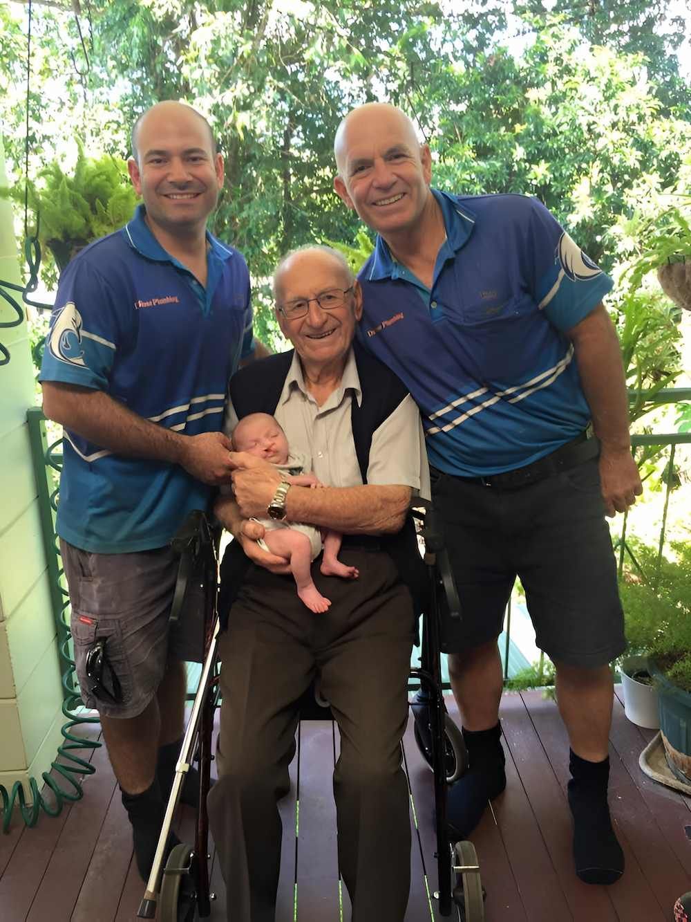 Three Men Are Standing Around an Older Man in a Wheelchair Holding a Baby — De Rose Plumbing In Bayview Heights, QLD