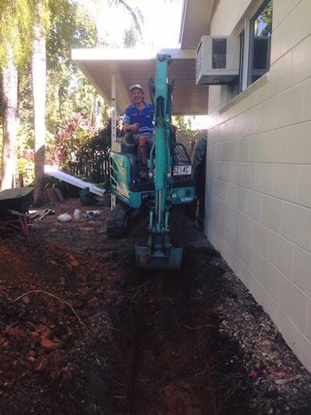 A Man is Driving a Small Excavator in Front of a House — De Rose Plumbing In Bayview Heights, QLD