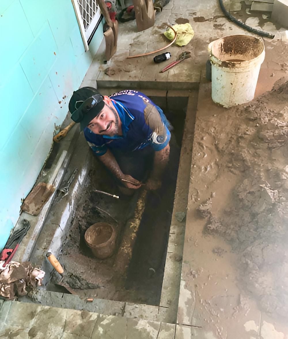 A Man is Kneeling in a Hole in the Ground Looking at a Pipe — De Rose Plumbing In Bayview Heights, QLD