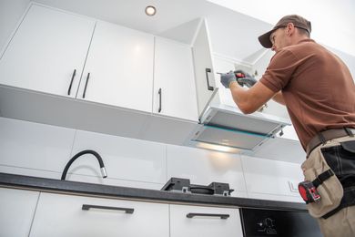 Une personne installe une hotte aspirante dans une cuisine blanche, à l'aide d'un tournevis.