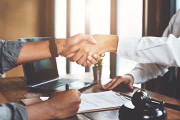 Two people shaking hands across a desk with a laptop and gavel in a legal office
