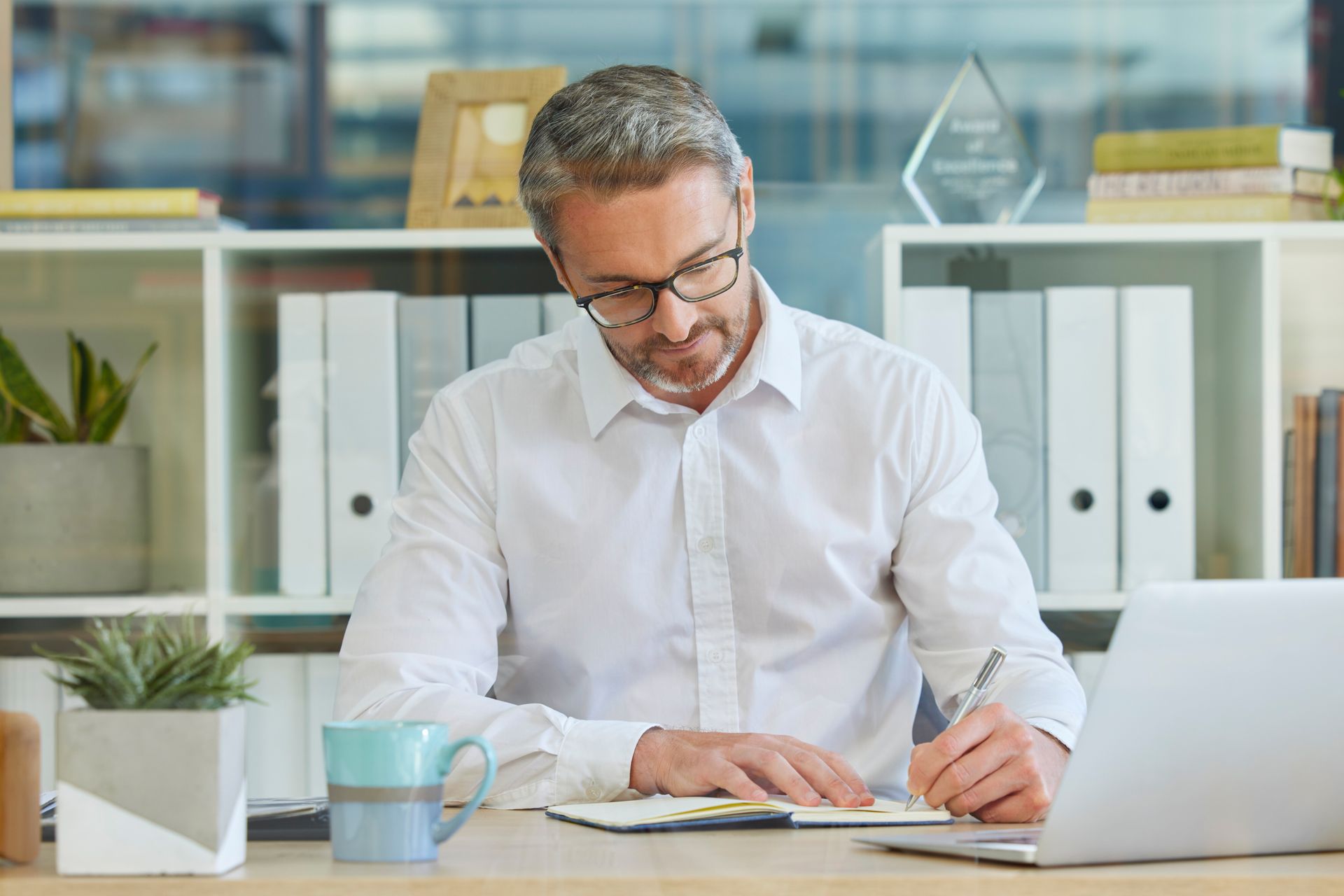 Man in a white shirt writing at a desk with a laptop in a bright office