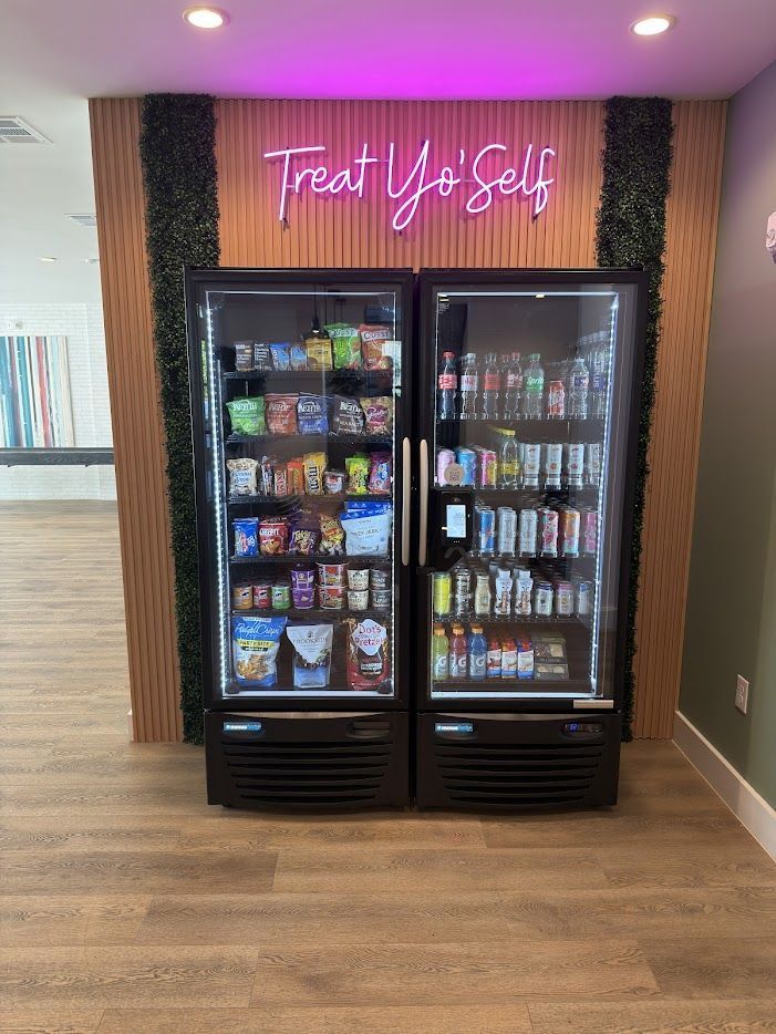 Snack and drink vending machines under a pink neon “Treat Yo’ Self” sign in a hallway