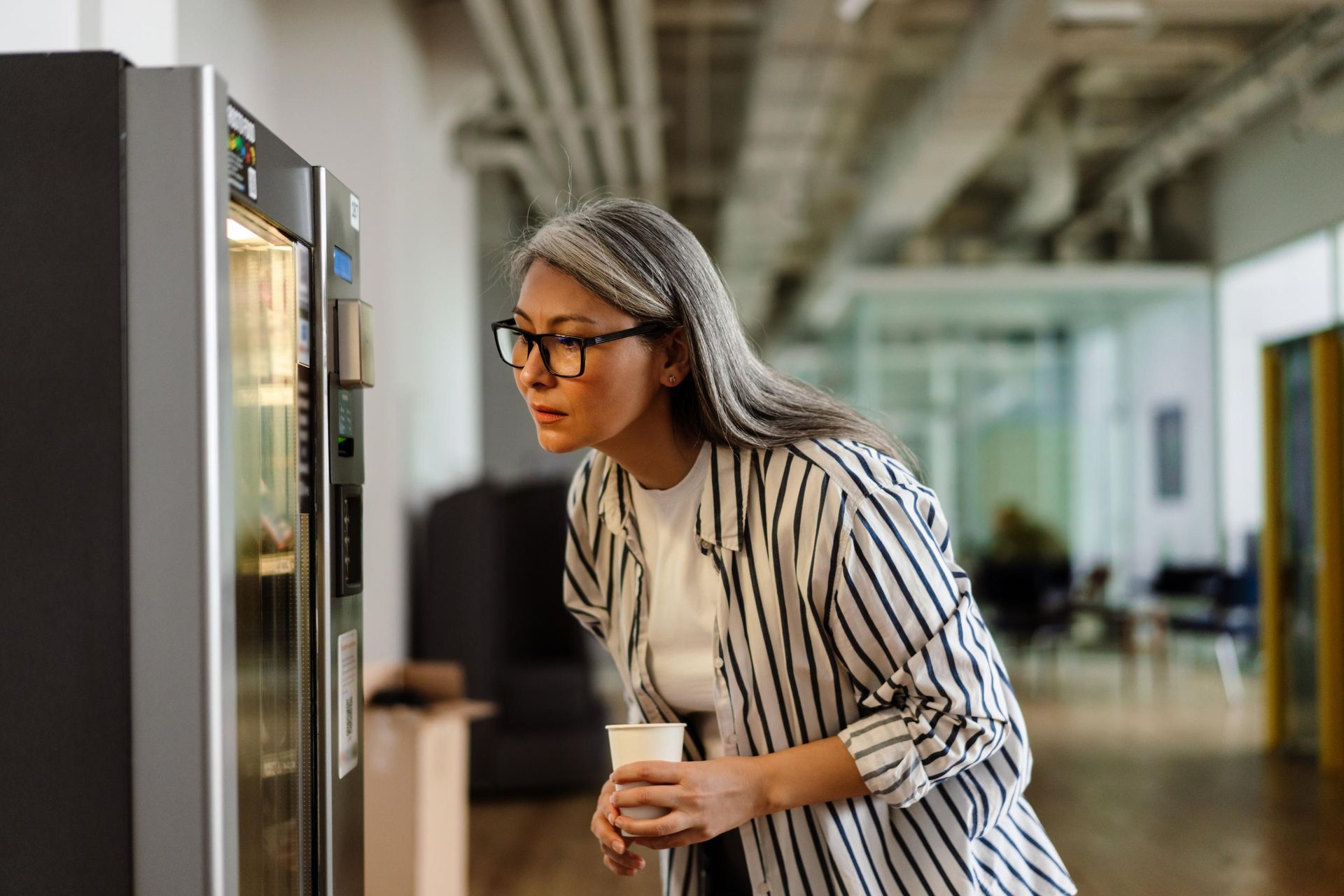 Woman with glasses looking at a vending machine, holding a cup in an office setting.