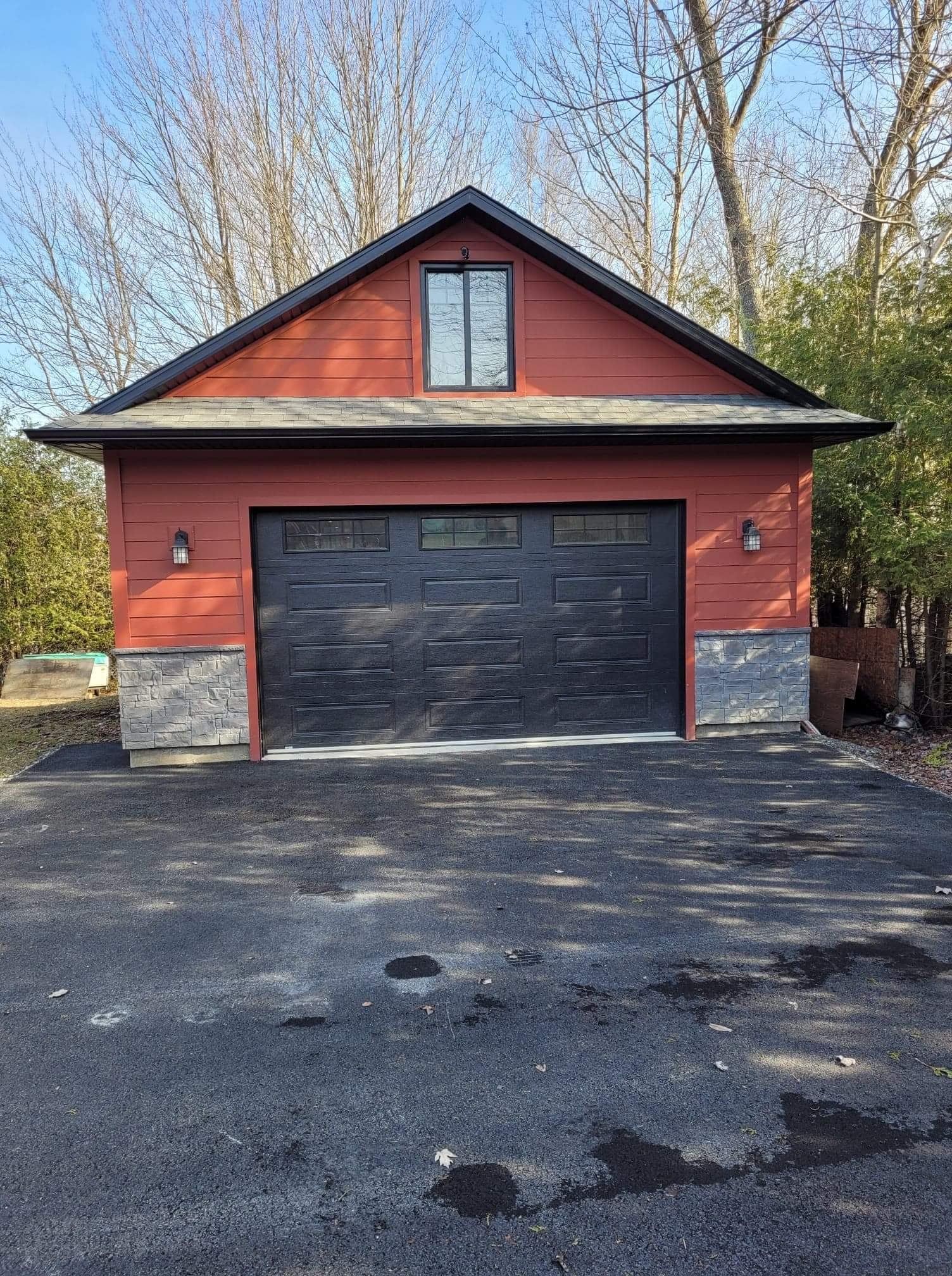 Garage rouge avec porte noire, soubassement en pierre et petite fenêtre, donnant sur une allée pavée.