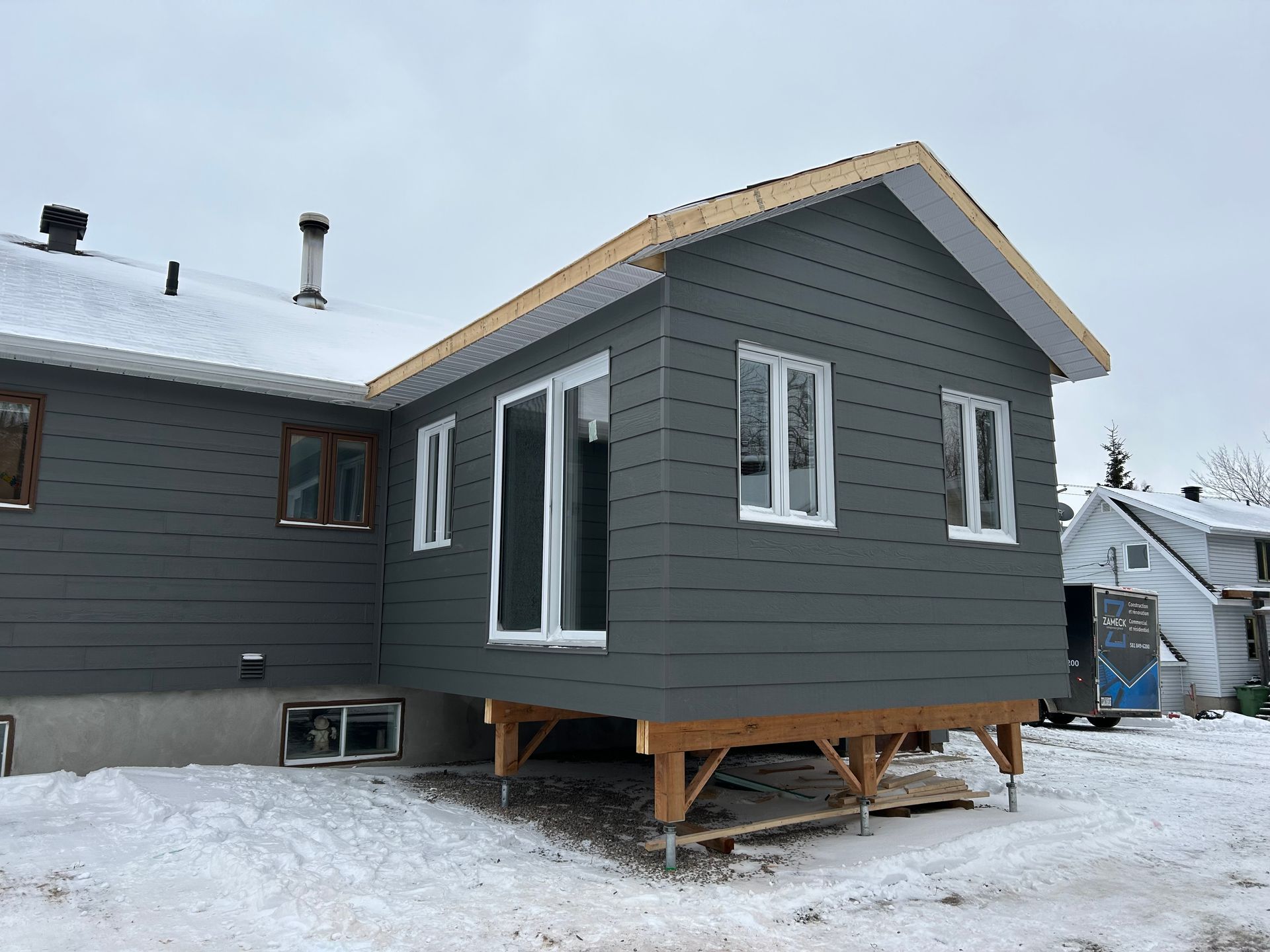 Extension de maison grise avec supports en bois, fenêtres et bardage, par une journée enneigée.